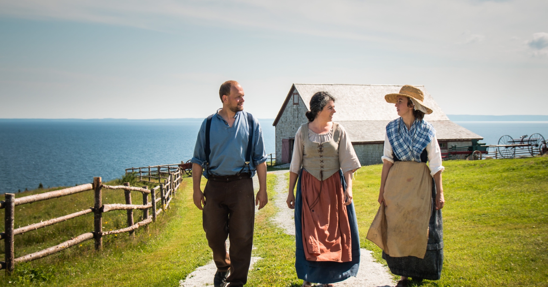 Three people in period clothing walk outside with the ocean in the background