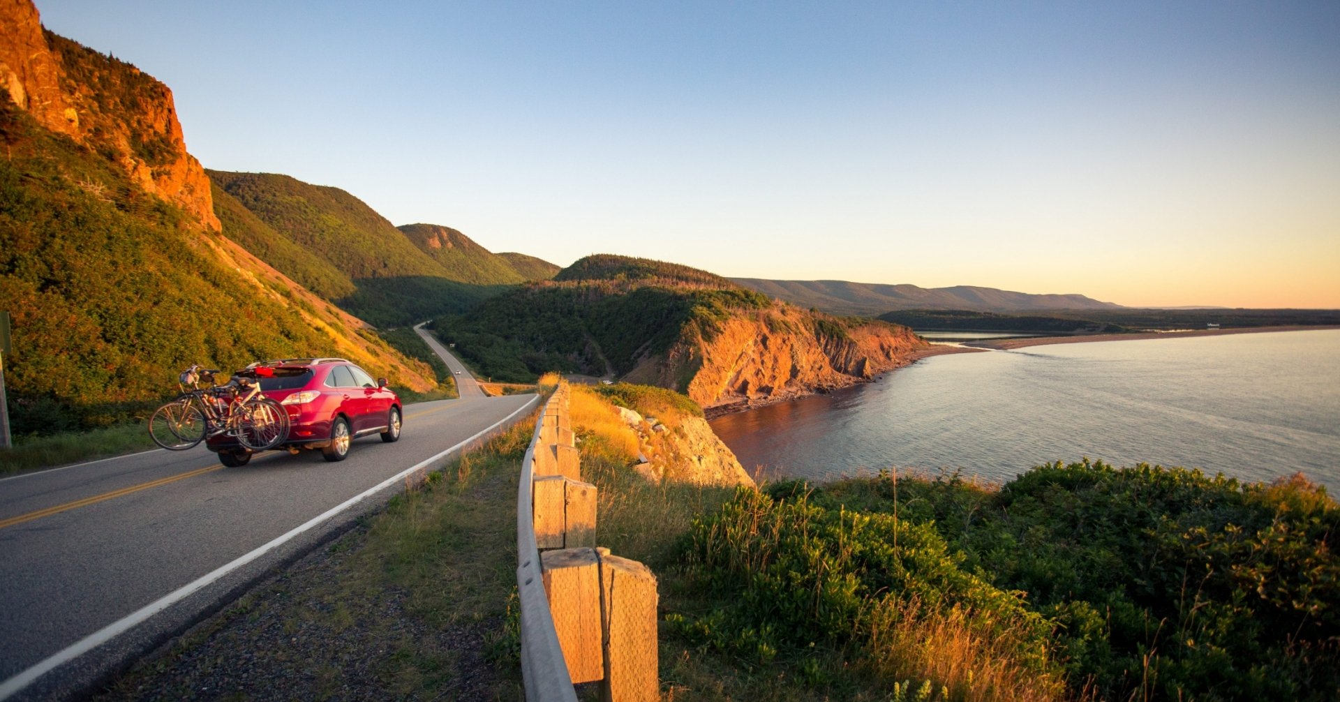 A red car with bikes attached drives down a highway overlooking the ocean